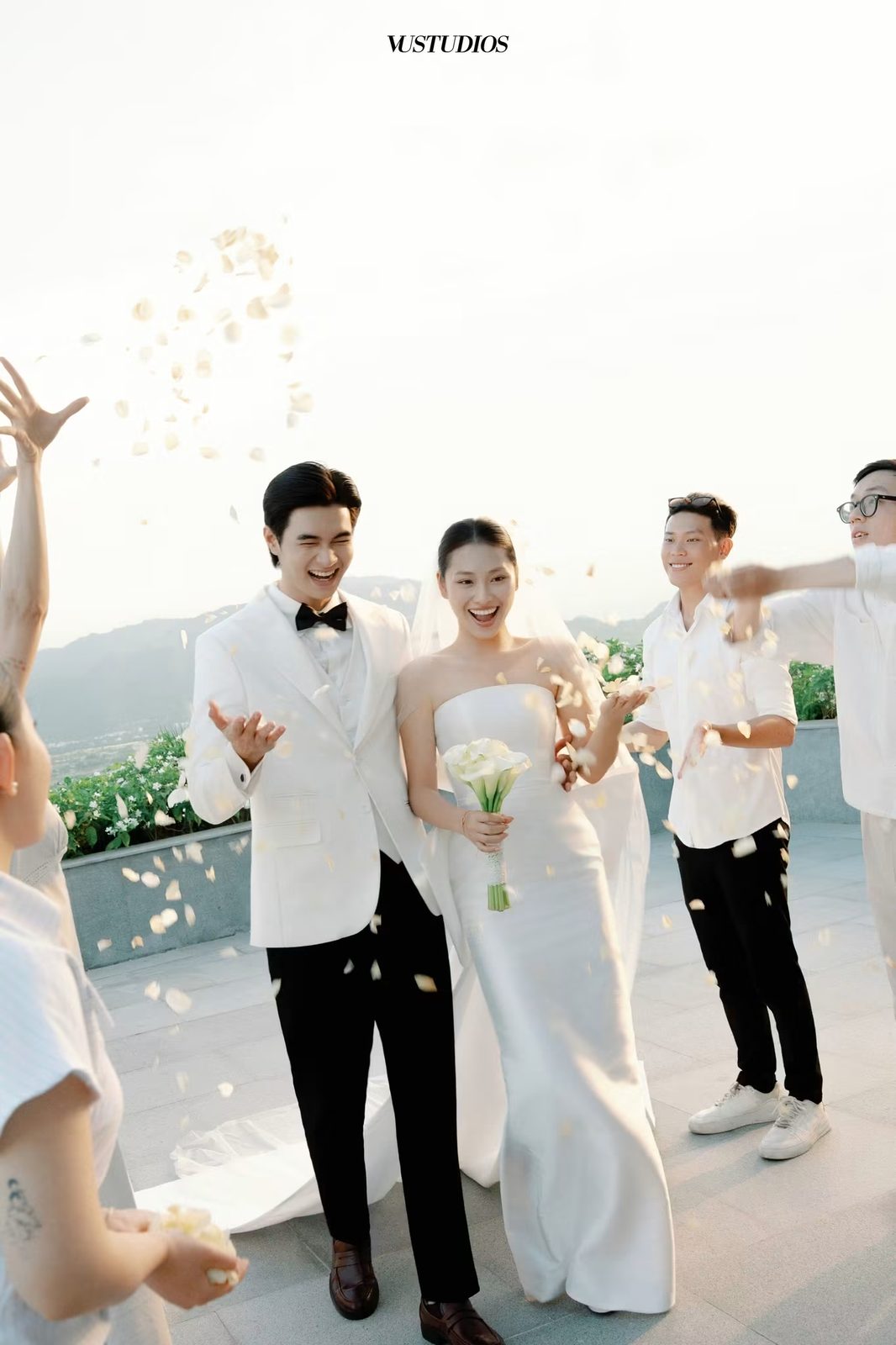 Chú rể suit trắng nơ đen và cô dâu váy trơn cầm hoa, cánh hoa tung trong tiệc ngoài trời