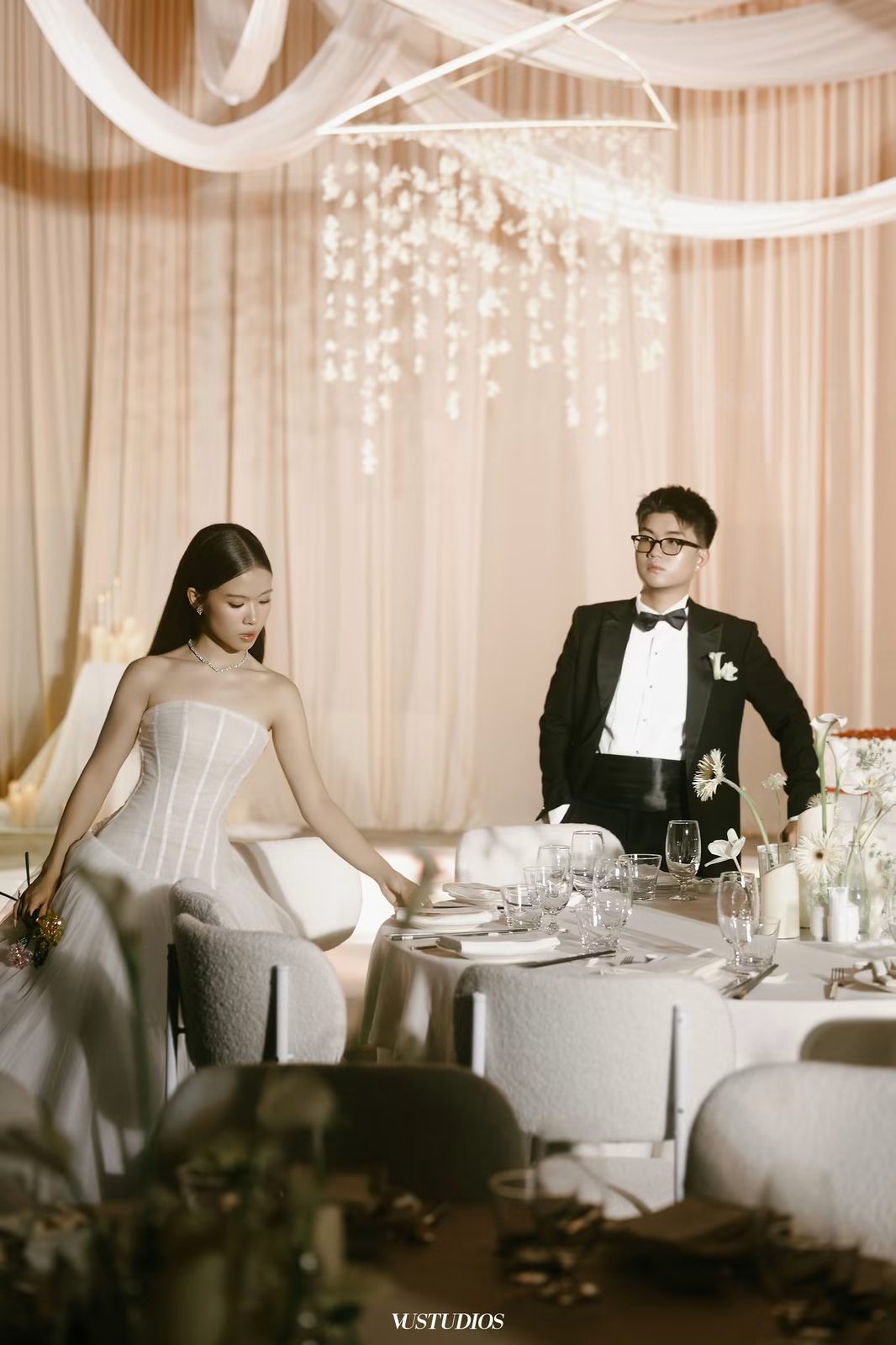 Chú rể tuxedo nơ đen và cô dâu váy trơn cúp ngực bên bàn tiệc rèm hồng, đèn pha lê