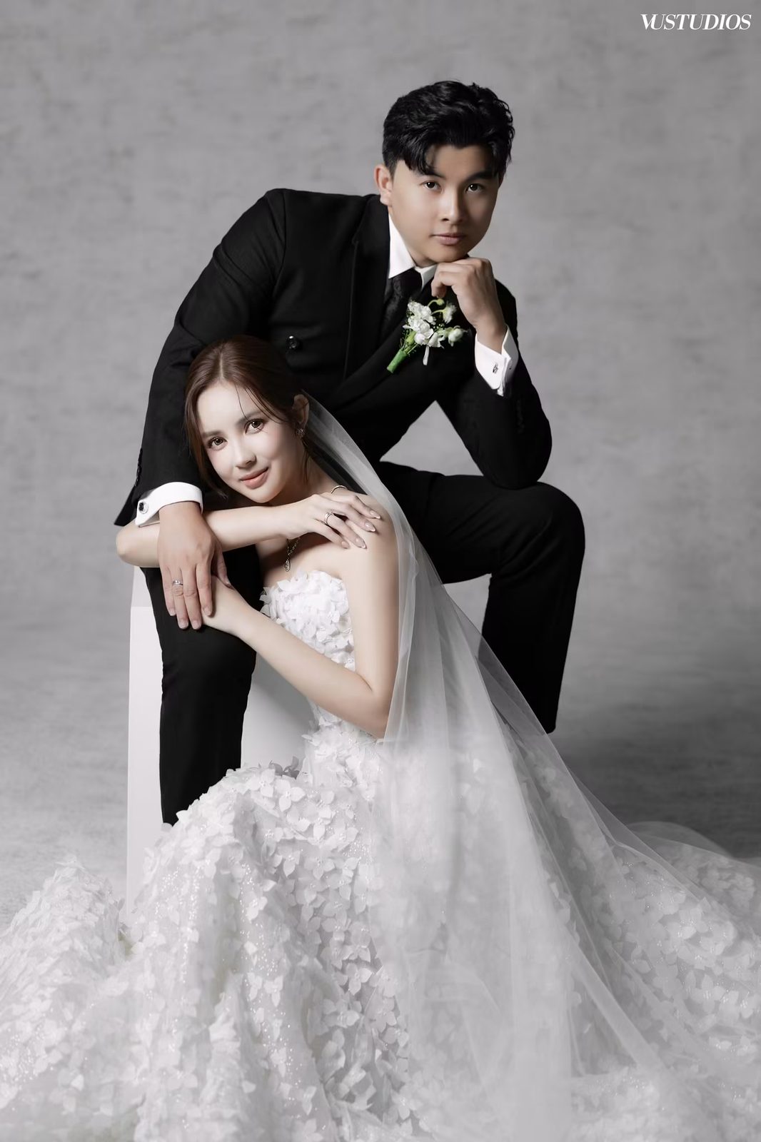 Cô dâu váy cưới xòe phủ hoa nổi và chú rể tuxedo đen tạo dáng bên nhau trên nền xám