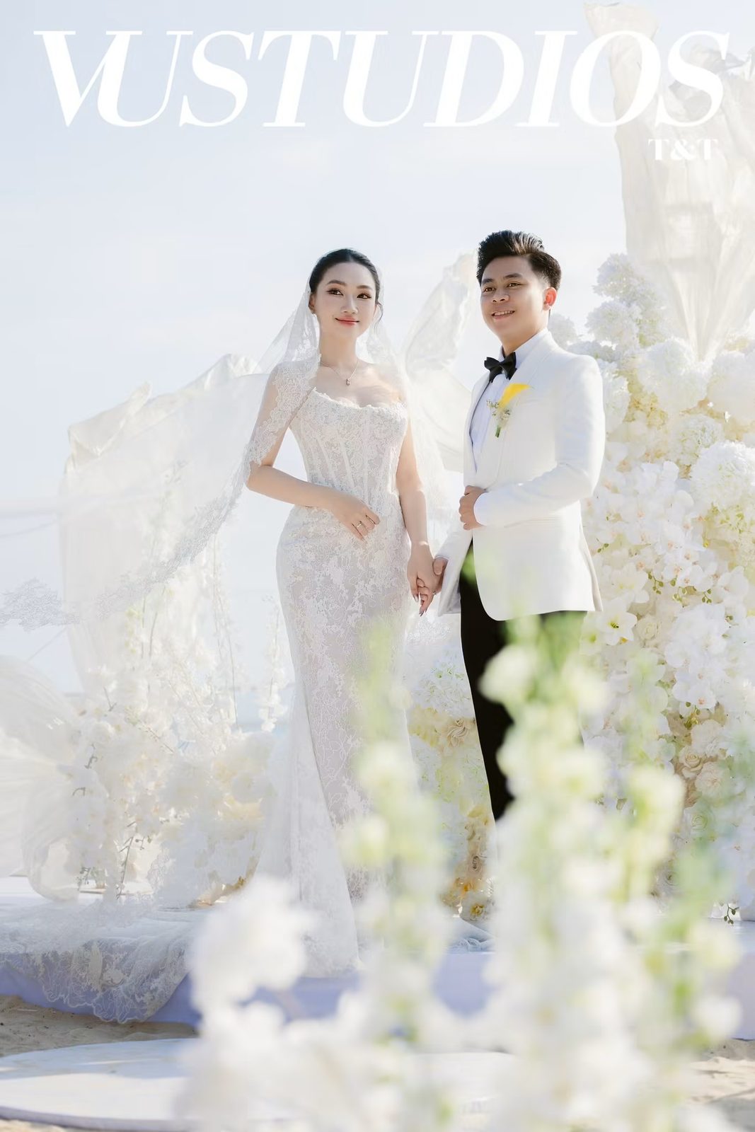 Cô dâu váy ren phủ voan và chú rể vest trắng nắm tay giữa hoa trắng trong tiệc ngoài trời
