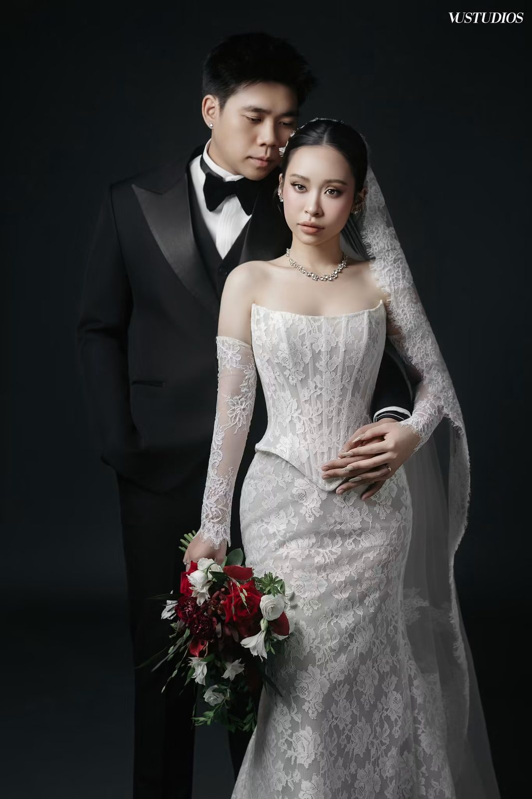 Chú rể tuxedo ve lụa nơ đen ôm cô dâu váy ren cúp ngực tay dài, cầm hoa đỏ trên nền tối