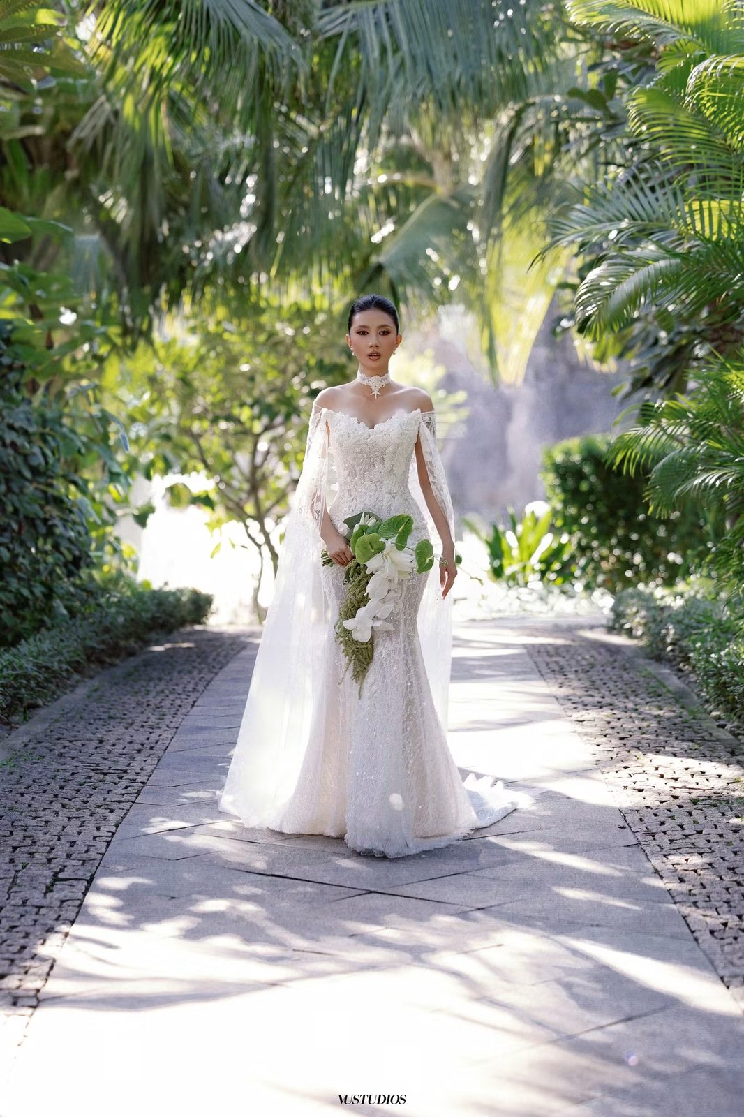 Cô dâu váy cưới đuôi cá ren kèm áo choàng cape bước trên lối đi trong khu vườn nhiệt đới