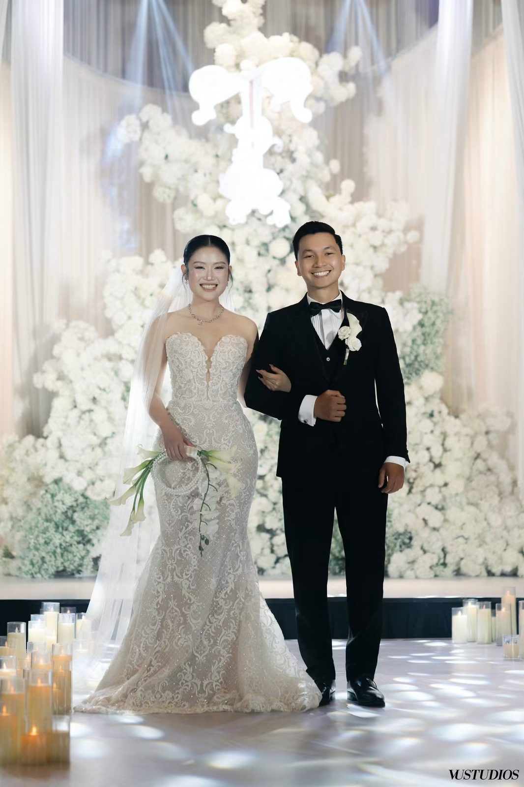 Cô dâu váy cưới đuôi cá ren và chú rể vest đen bước ra lễ đường hoa trắng, nến thắp sáng