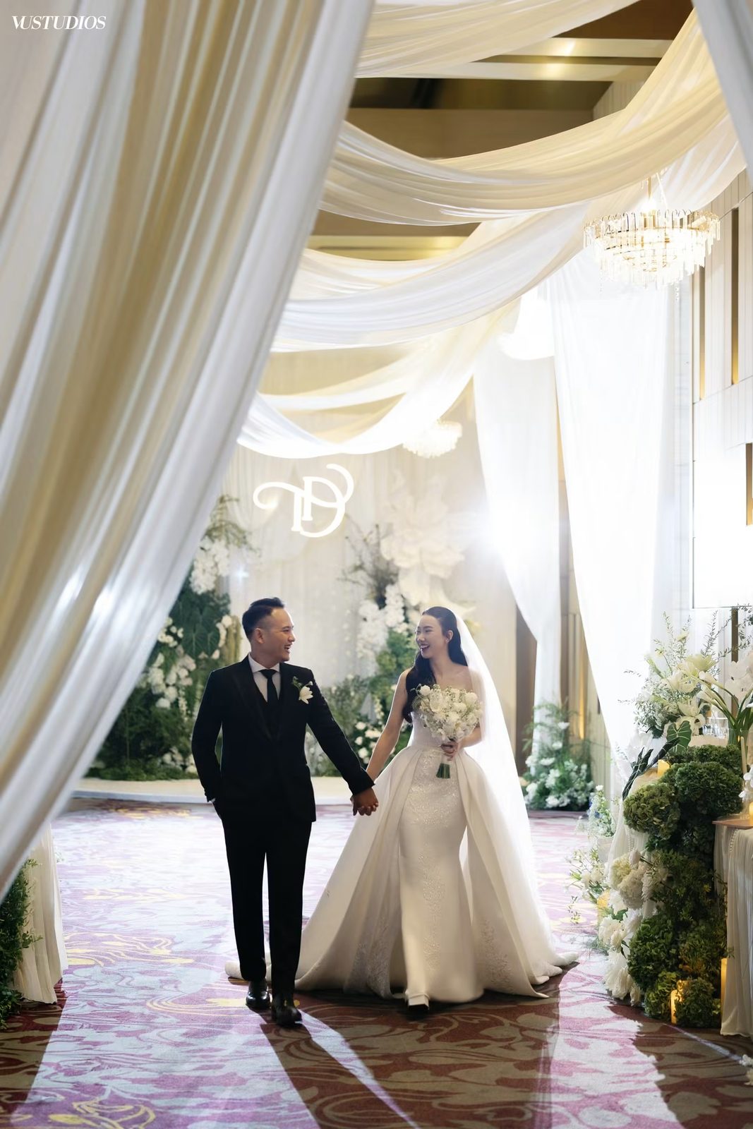 Cô dâu váy cưới đuôi cá kèm tùng ngoài tháo rời nắm tay chú rể trong sảnh tiệc rèm voan