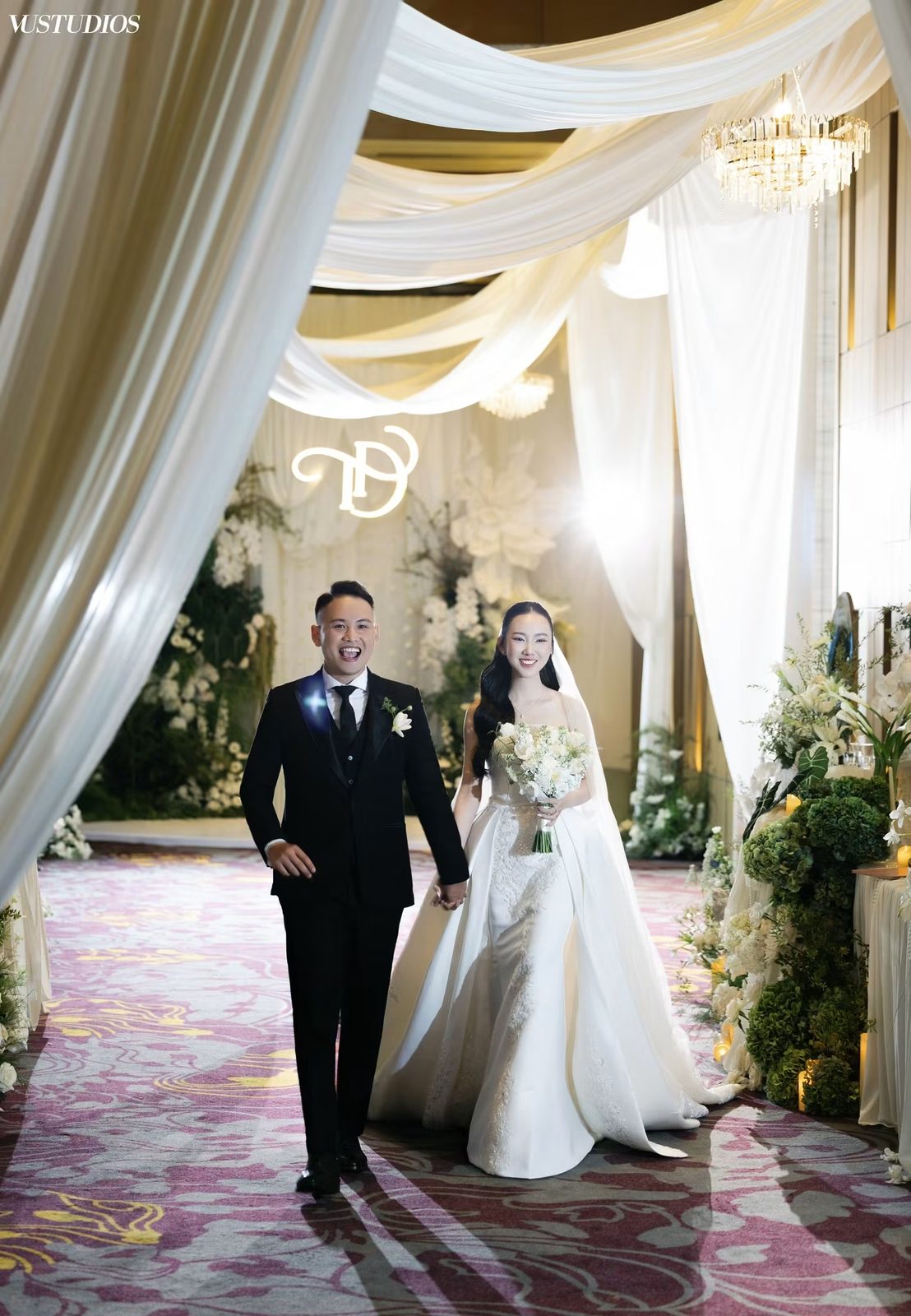 Cô dâu váy lụa satin trơn nắm tay chú rể vest đen bước trong sảnh tiệc rèm voan