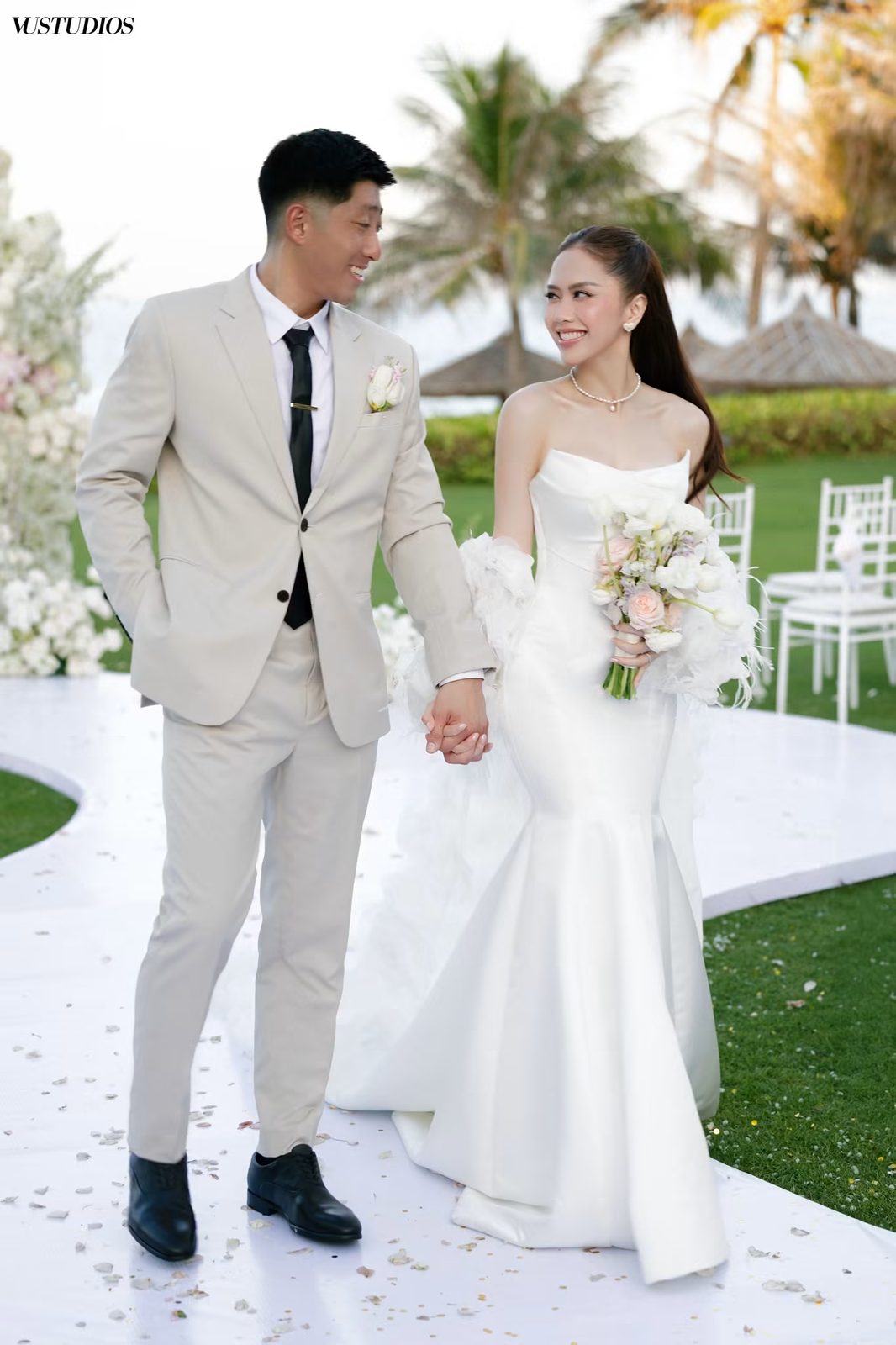 Cô dâu váy lụa satin đuôi cá nắm tay chú rể vest be trên lối đi trắng ngoài trời