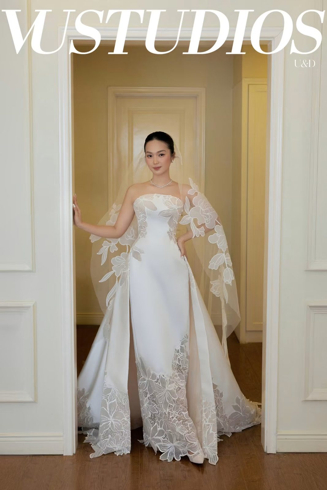 Cô dâu váy lụa satin cúp ngực với tùng ngoài ren tháo rời và voan đứng ở khung cửa trắng
