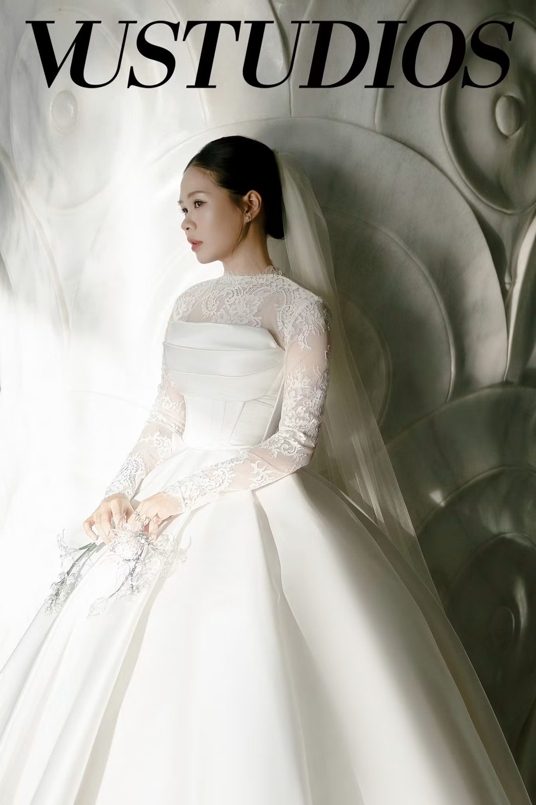 Cô dâu váy cưới tay ren cổ cao với chân váy lụa trơn xòe, ngồi nghiêng bên tường họa tiết