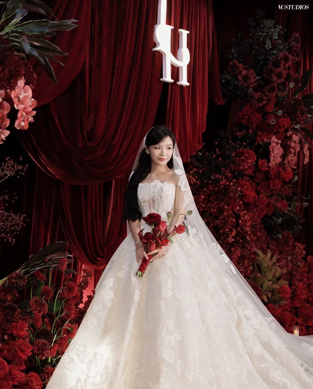 Cô dâu váy cưới xòe phủ ren hoa nổi bên phông rèm đỏ và hoa đỏ, tay cầm bó hoa đỏ