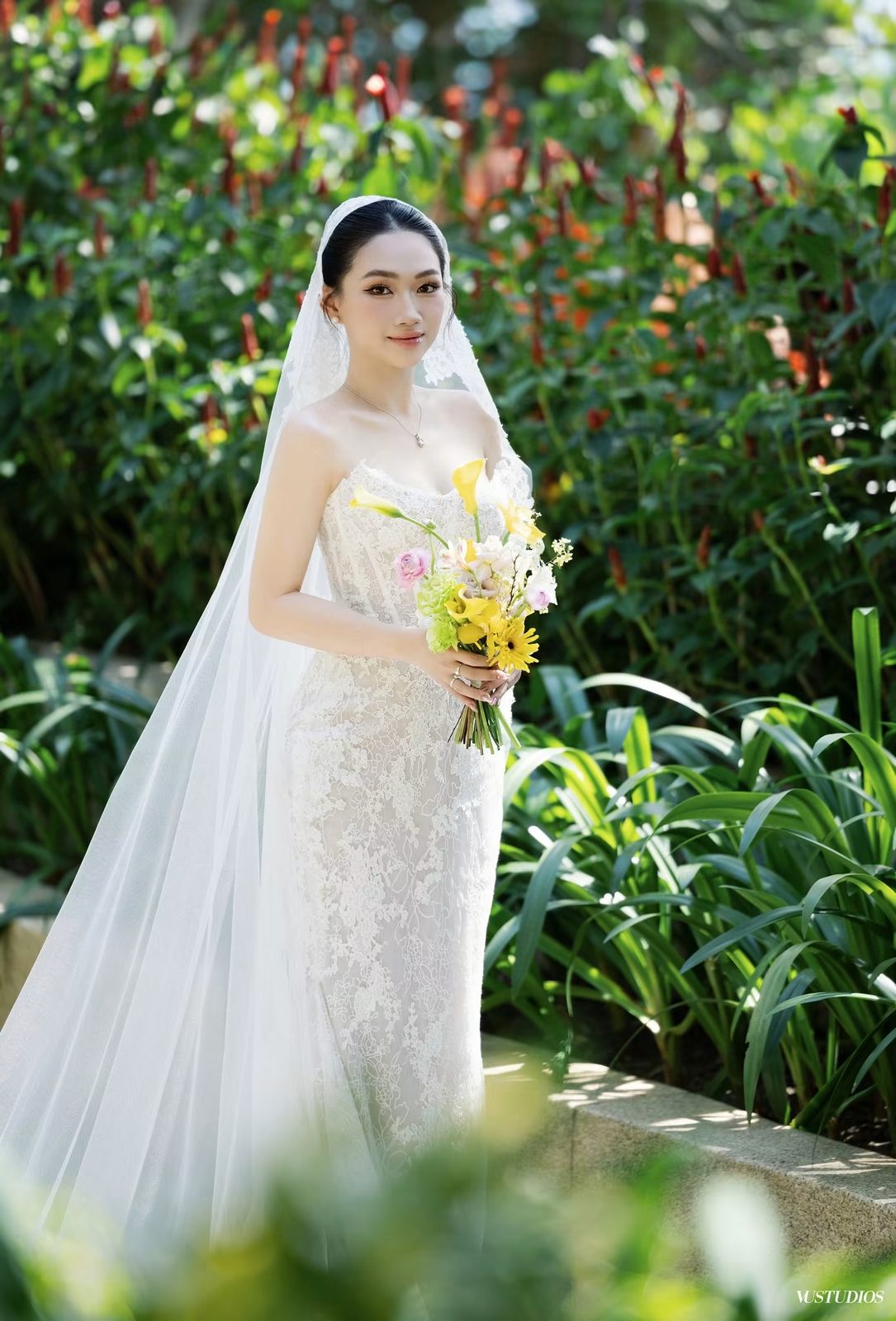 Cô dâu váy cưới ren Alençon cúp ngực với voan dài, cầm bó hoa vàng trong khu vườn xanh