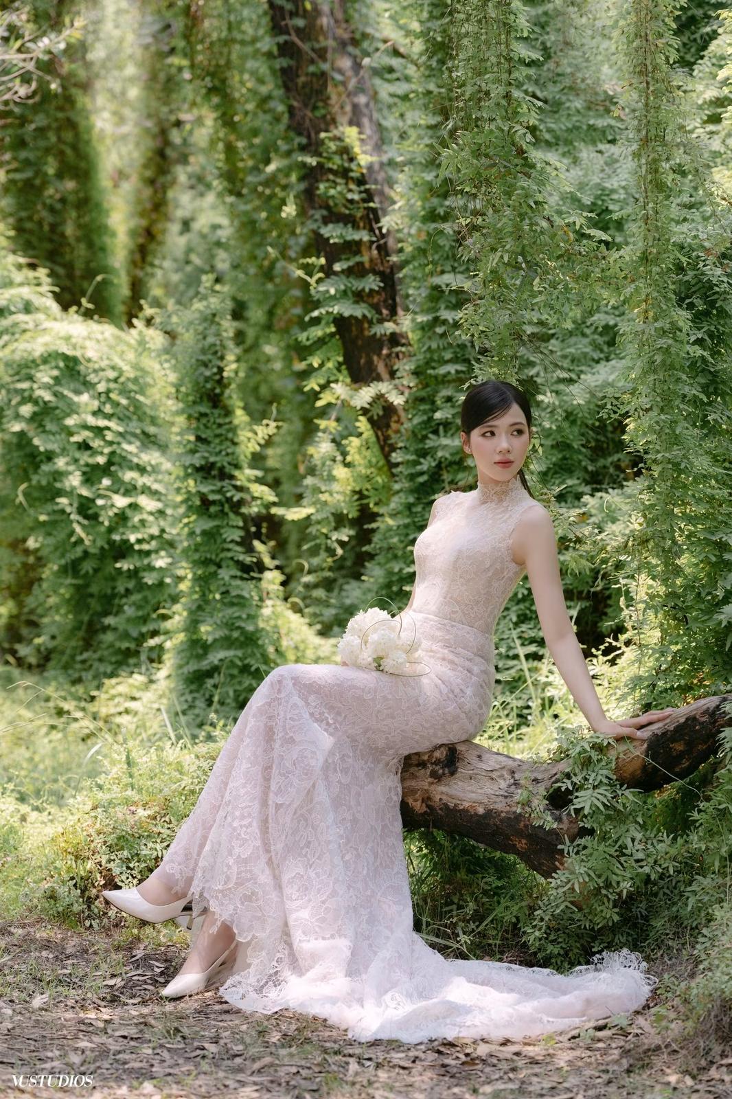 Cô dâu váy ren Chantilly nhẹ ngồi trên thân gỗ giữa khu rừng xanh, tay cầm hoa trắng