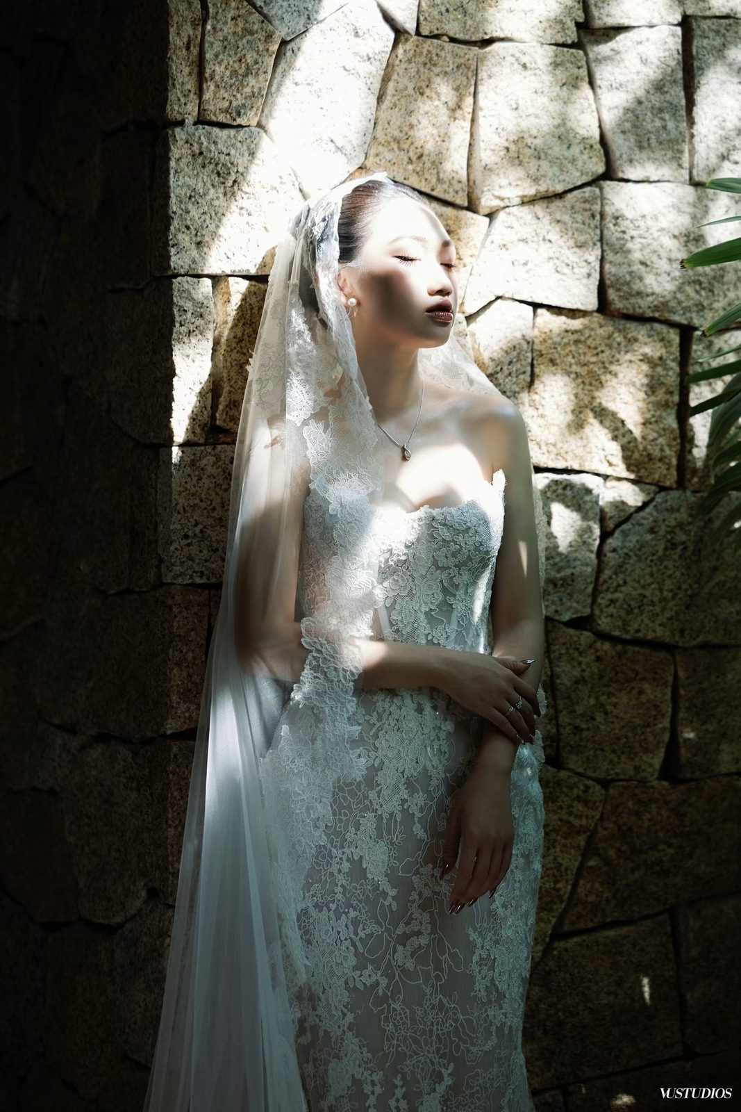 Cô dâu váy cưới ren mỏng phủ voan đứng nhắm mắt bên bức tường đá giữa ánh sáng và bóng đổ