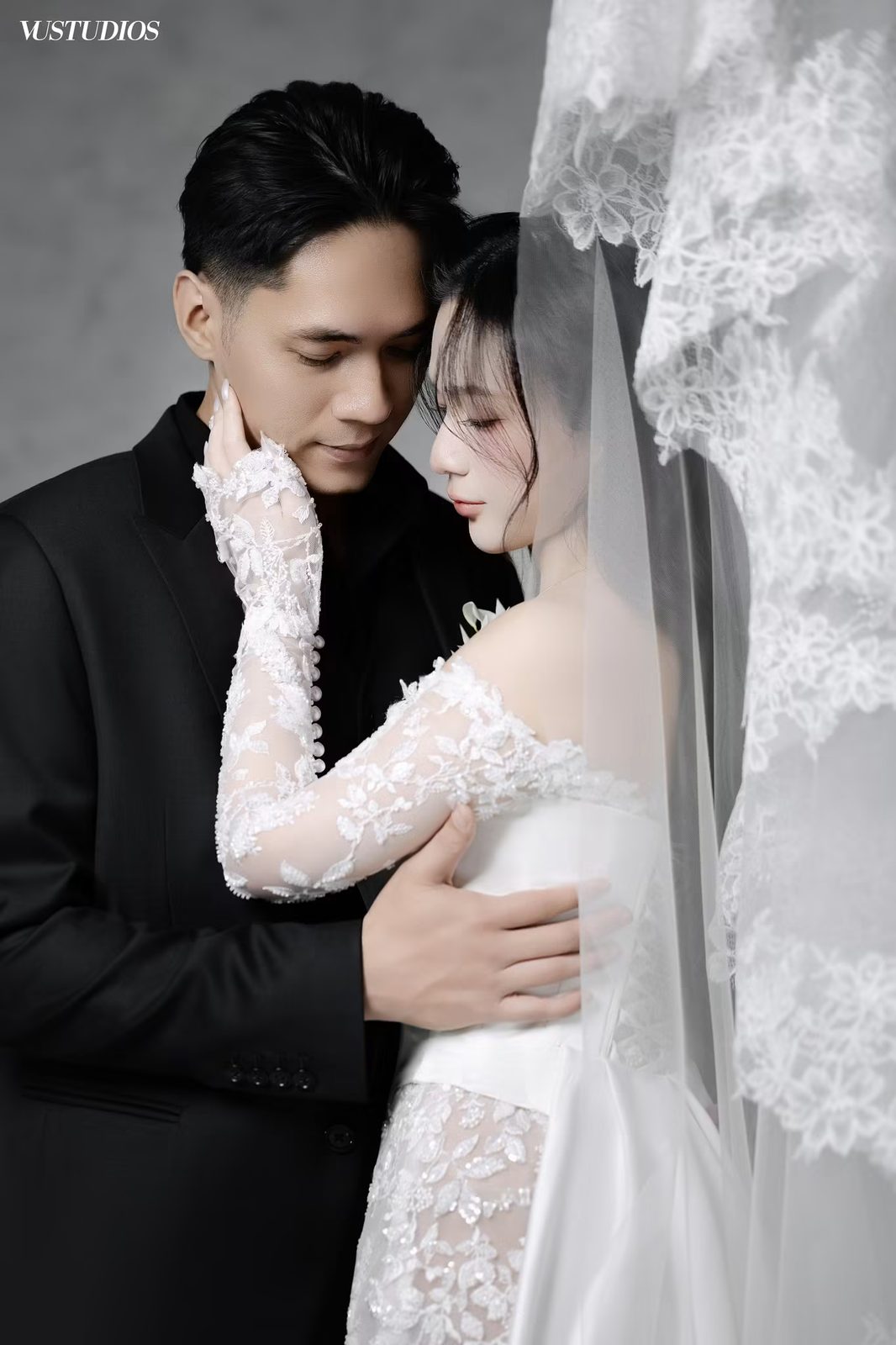 Cô dâu váy ren tay dài tựa vào chú rể vest đen, voan ren viền hoa phủ phía sau
