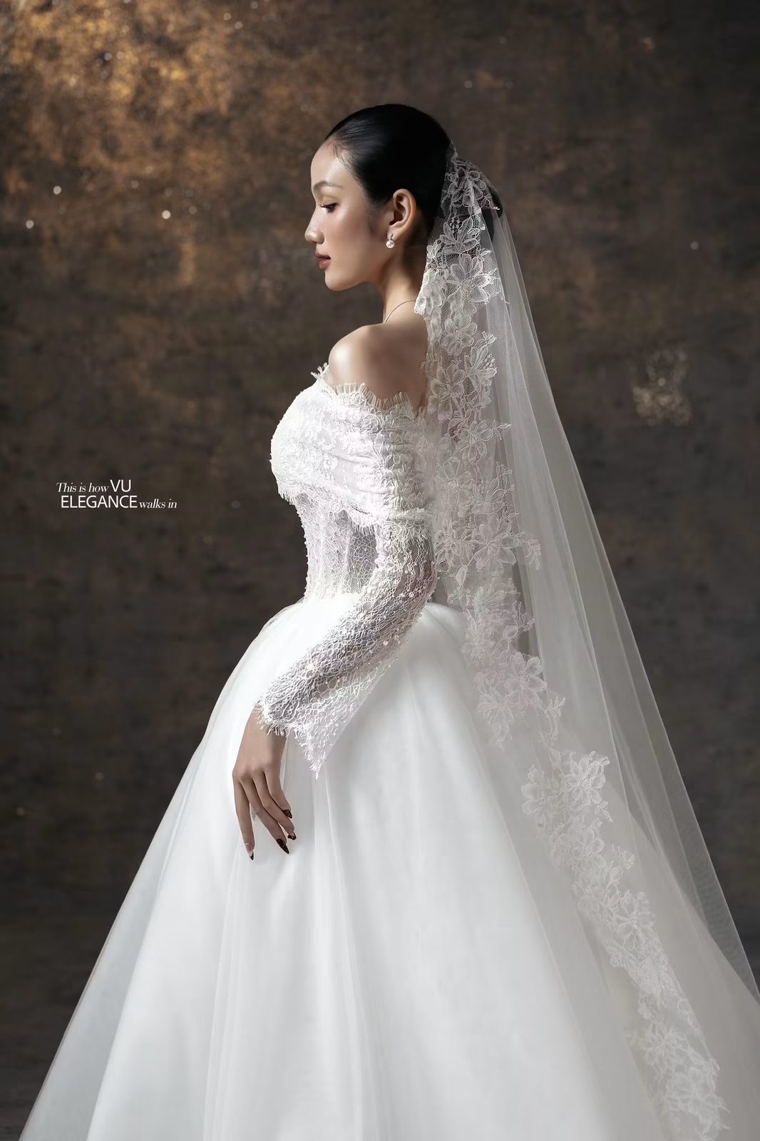 Người mẫu nghiêng mặt trong váy cưới ren tay dài, chân váy tulle xòe và voan viền ren