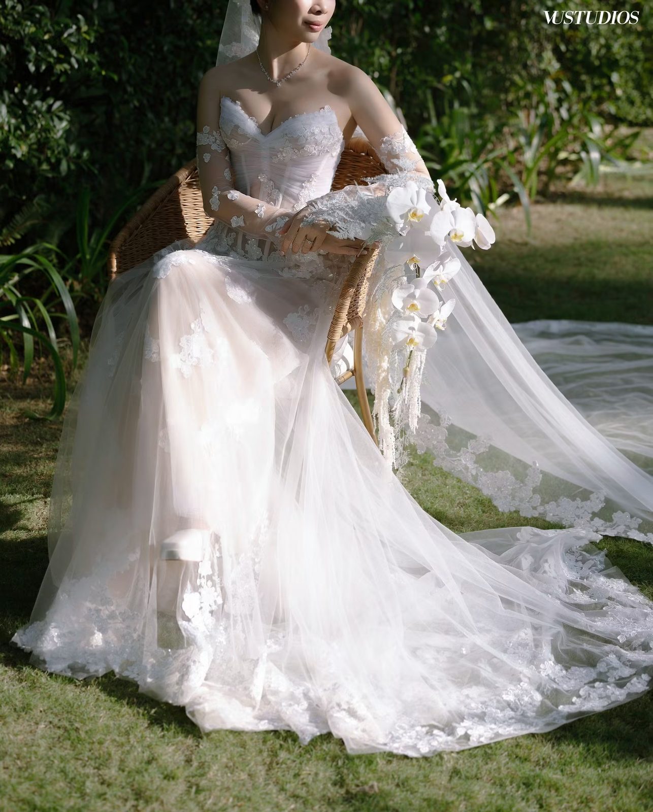 Cô dâu váy ren thêu chìm tùng tulle dài ngồi trên ghế mây ngoài sân cỏ, cầm hoa lan trắng