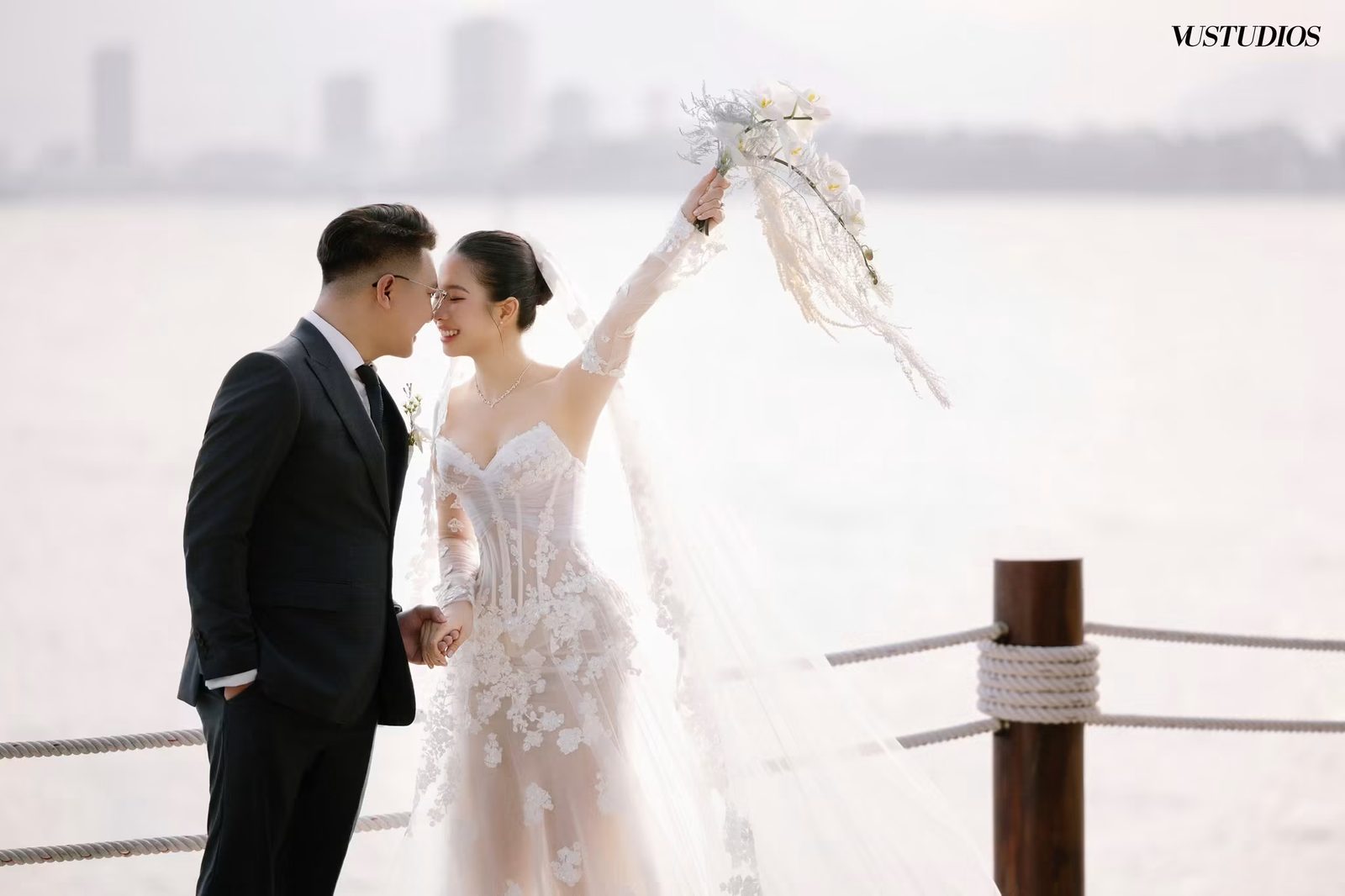 Cô dâu váy ren thêu hoa nổi và chú rể vest đen trao nhau cái hôn bên lan can bờ sông