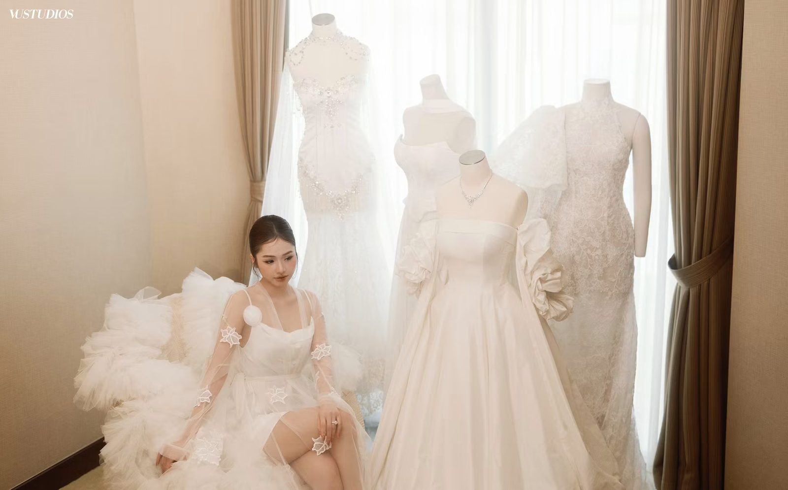 Cô dâu ngồi giữa các mẫu váy cưới trên ma-nơ-canh trong showroom bên cửa sổ rèm voan