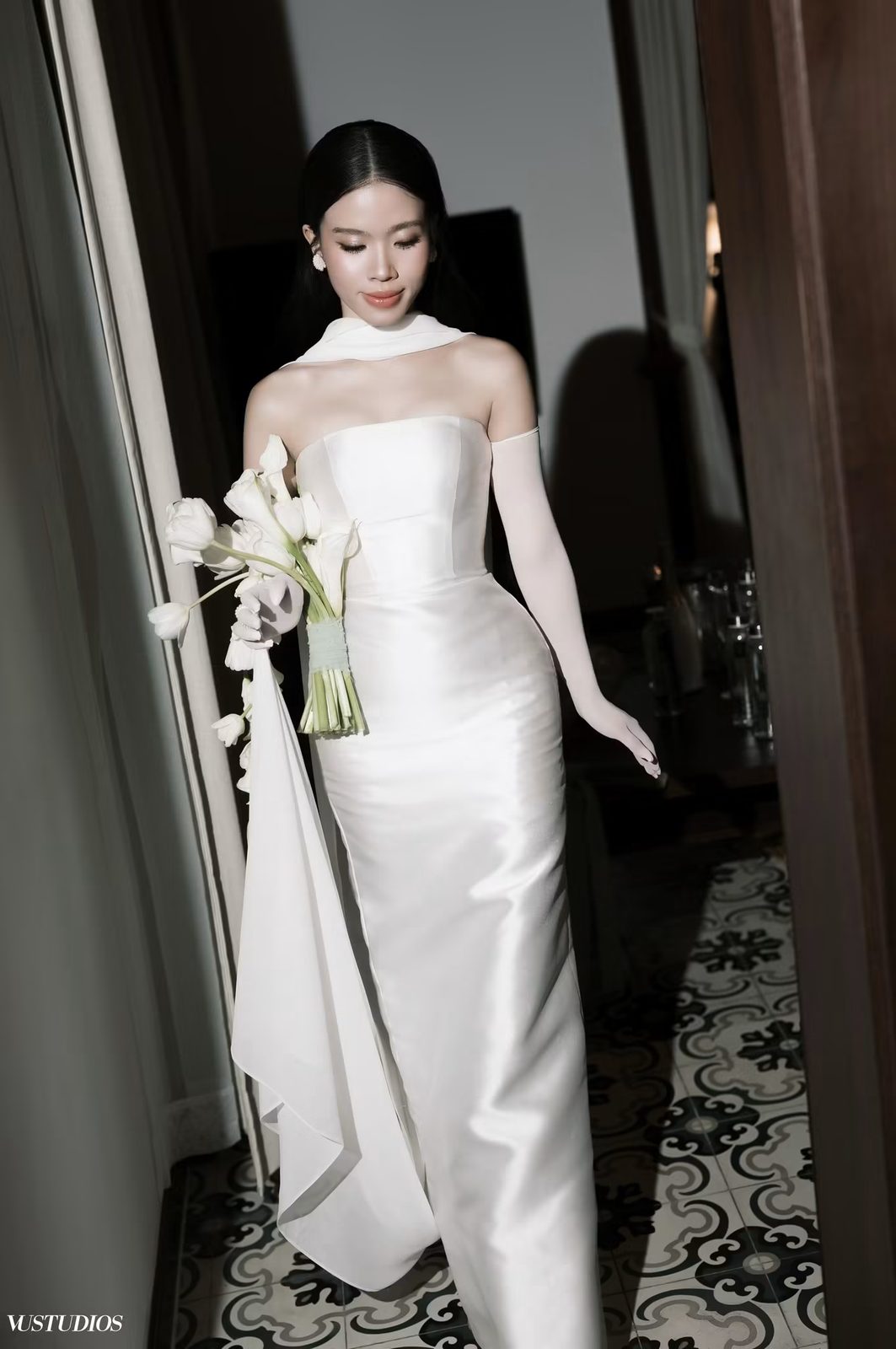 Cô dâu váy cưới trơn satin cúp ngực với găng tay dài và bó hoa cala trong hành lang tối