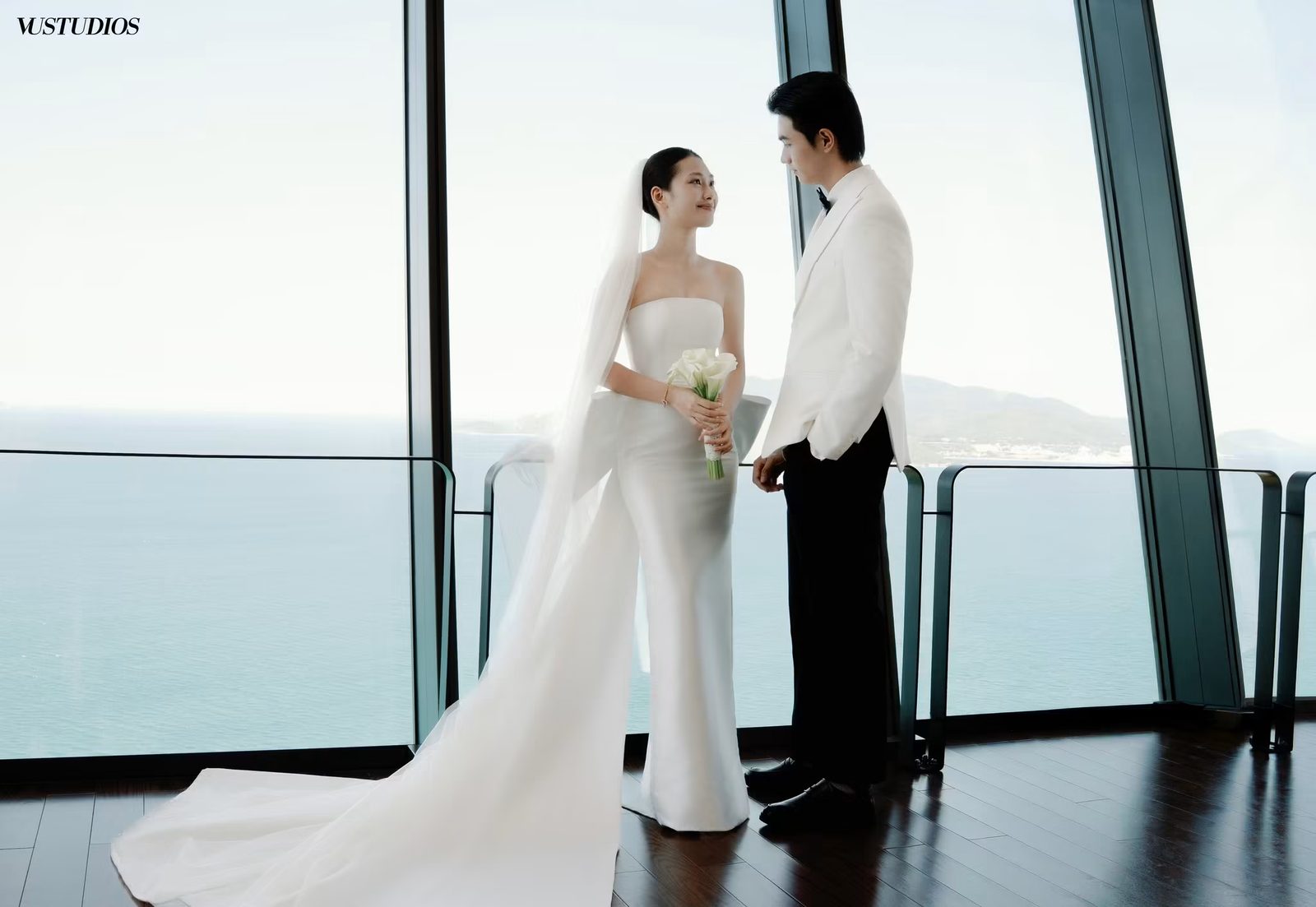 Cô dâu váy cưới trắng tinh đuôi dài và chú rể đứng bên cửa kính nhìn ra biển