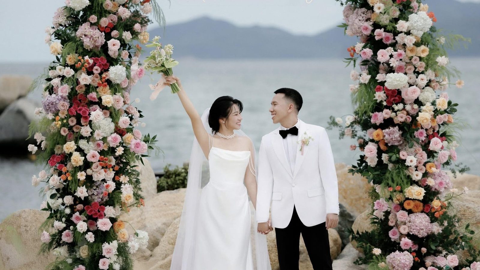 Cô dâu váy cưới trơn và chú rể vest trắng nắm tay trước cổng hoa bên bờ biển