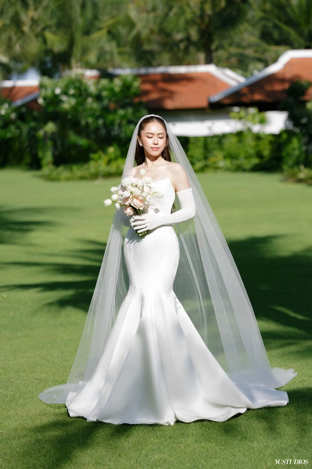 Cô dâu váy cưới trơn đuôi cá cầm hoa trên bãi cỏ, đeo găng tay dài và voan