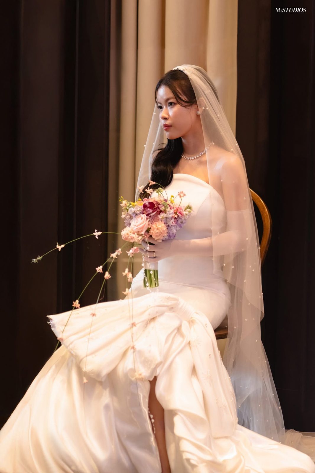 Cô dâu váy cưới trơn đuôi cá xếp dún và xẻ tà, phủ voan, ngồi cầm bó hoa
