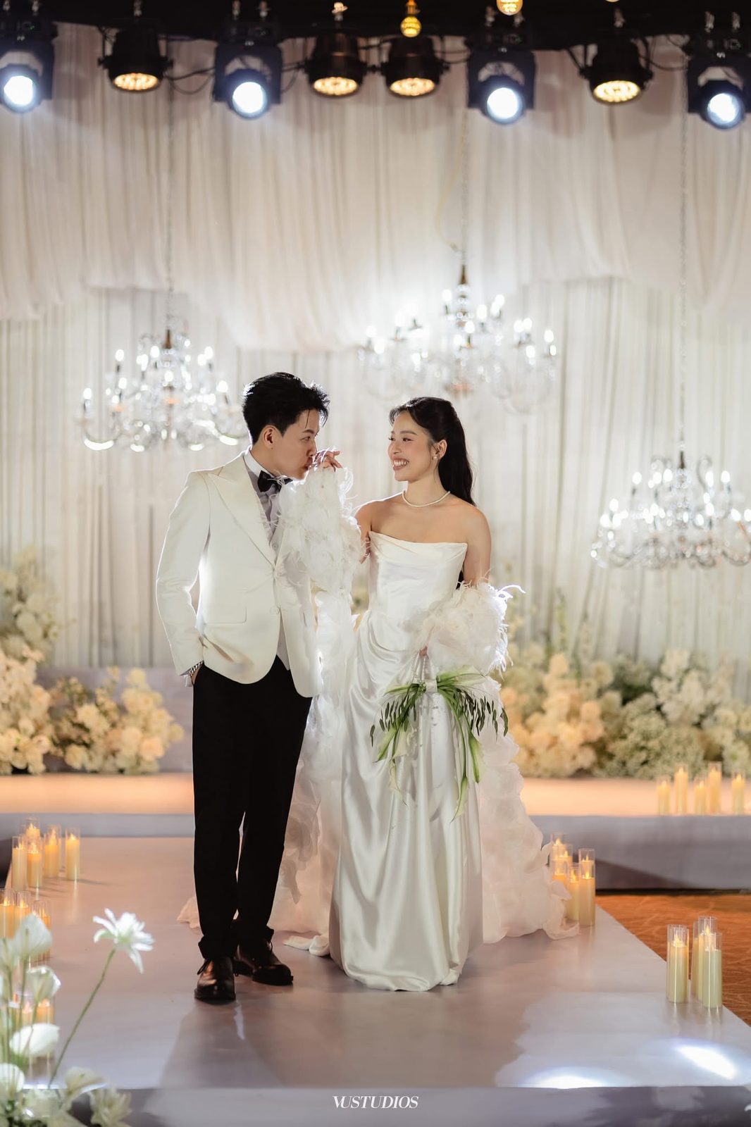 Cô dâu váy cưới trơn satin cúp ngực và chú rể vest trắng trên lối đi nến, đèn chùm pha lê